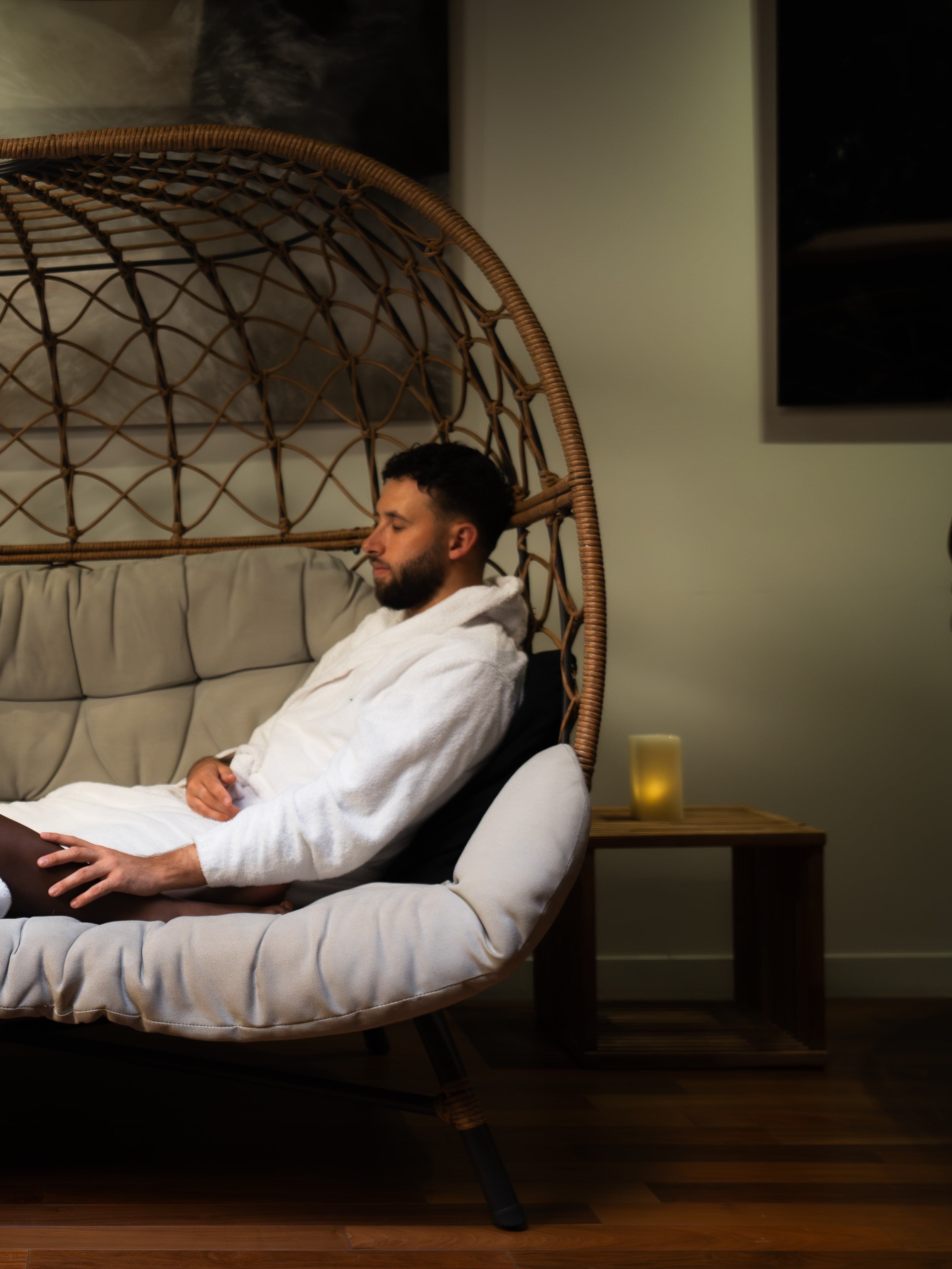 A couple in white bathrobes relaxing on a comfortable double chair. In the background, there is a small wooden table with a candle.