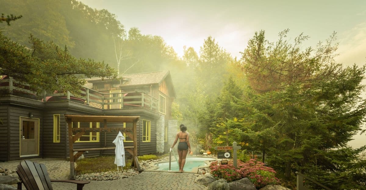 A person in swimwear stands at the edge of a hot tub, surrounded by lush trees and mist. A rustic wooden cabin is in the background. The setting conveys tranquility and nature.