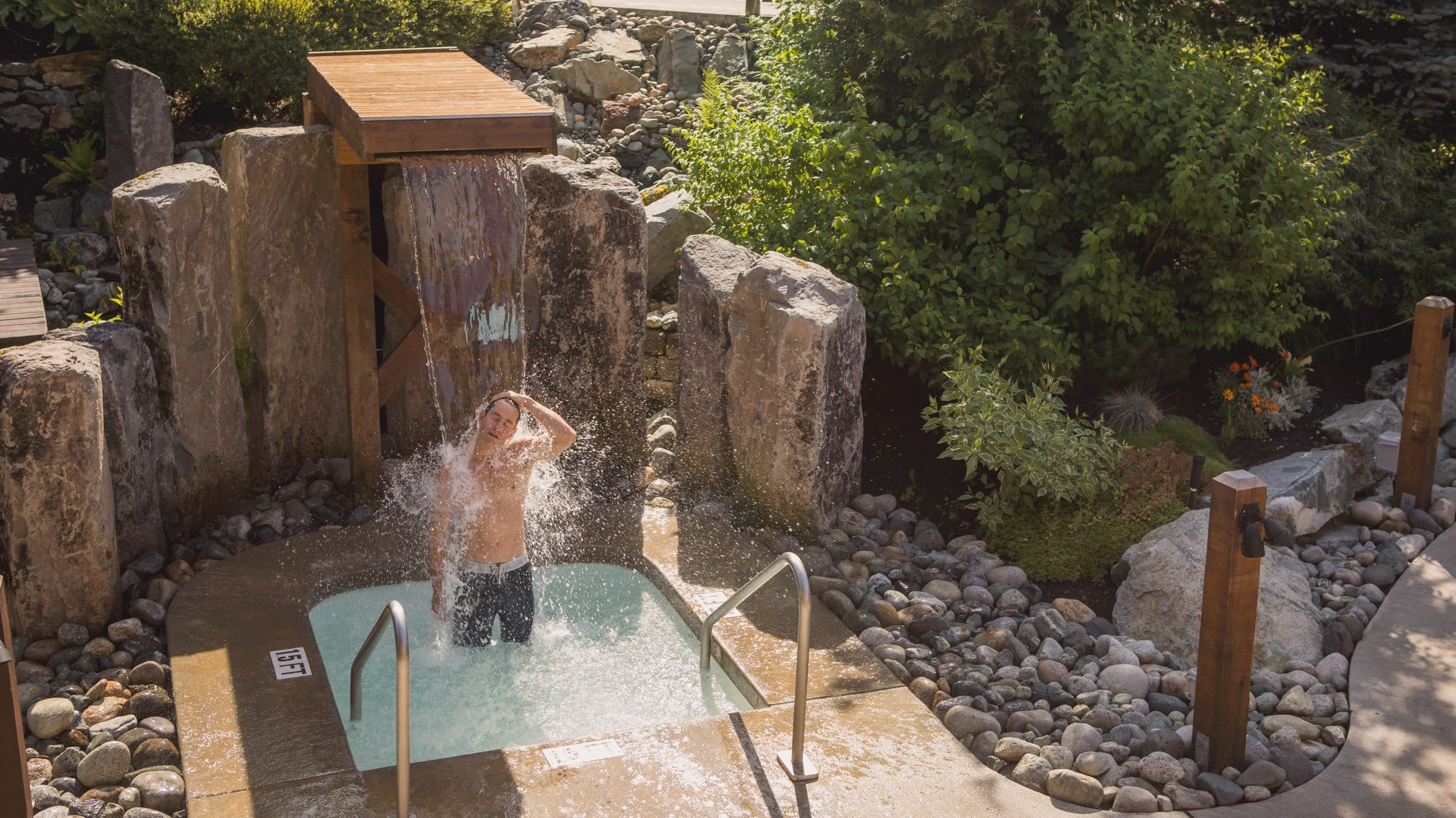 A man stretching his neck under the Nordic waterfall at Scandianve Spa Whistler and enjoying the summer sun rays.