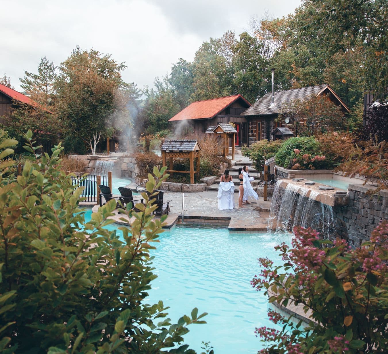 Outdoor spa scene featuring turquoise hot pools, a stone waterfall, and two guests in white robes walking along the pathway surrounded by lush garden landscaping and rustic wooden buildings.