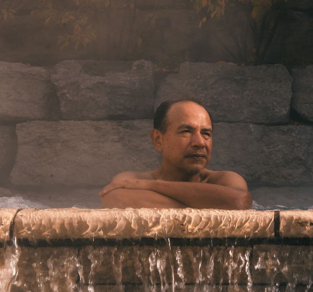 A man relaxing in an outdoor hot tub, leaning against the stone overflow with water flowing and steam all around. He is enjoying the soothing silence that reigns at Scandinave Spa.
