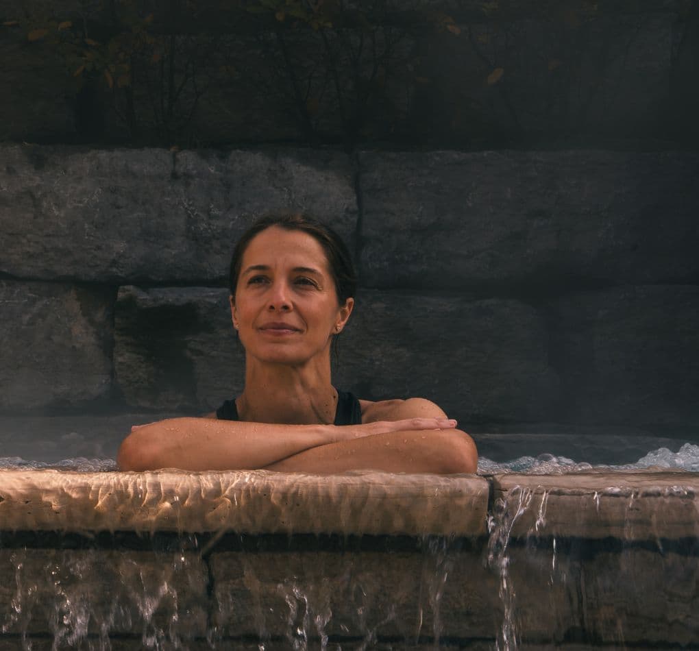 A woman relaxing in an outdoor hot tub, leaning against the stone overflow with water flowing and steam all around. She is enjoying the moment away from screens and digital devices at Scandinave Spa.
