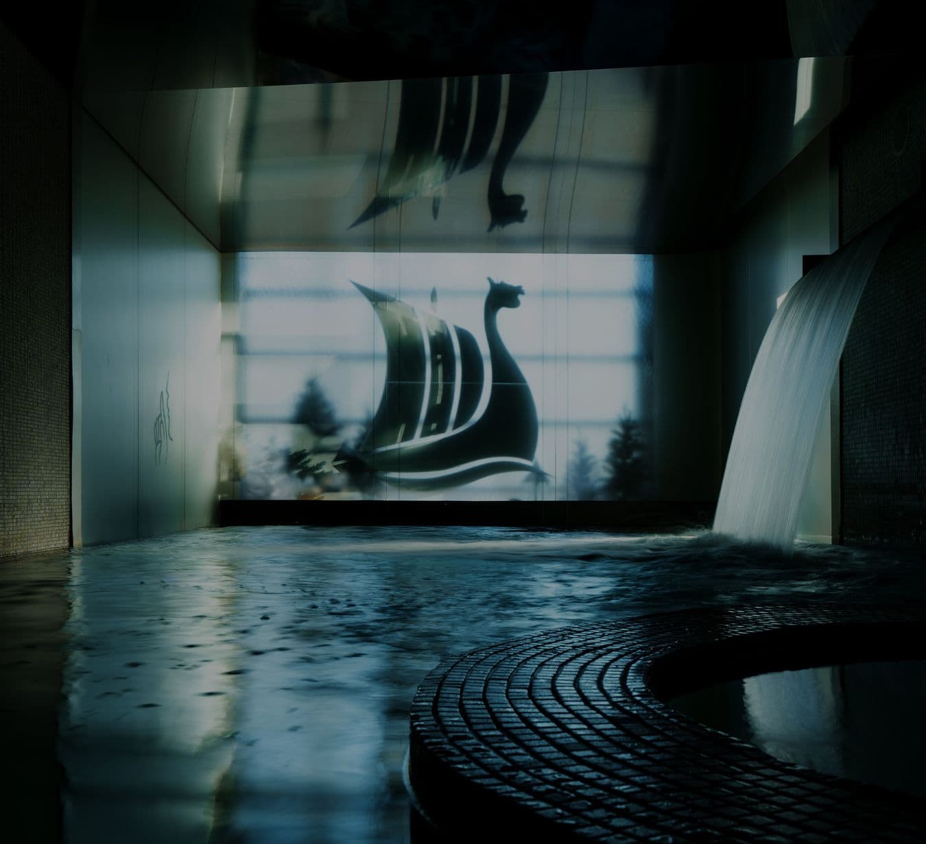 Piscine intérieure avec une chute thermale au premier plan. À l’arrière-plan, un mur illuminé présente le Drakkar, logo Scandinave Spa, au design de bateau viking stylisé, créant une ambiance apaisante.