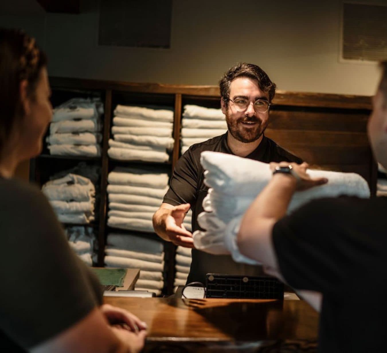 Kind-looking customer service agent handing folded towels to customers at a reception counter with shelves filled with towels in the background.