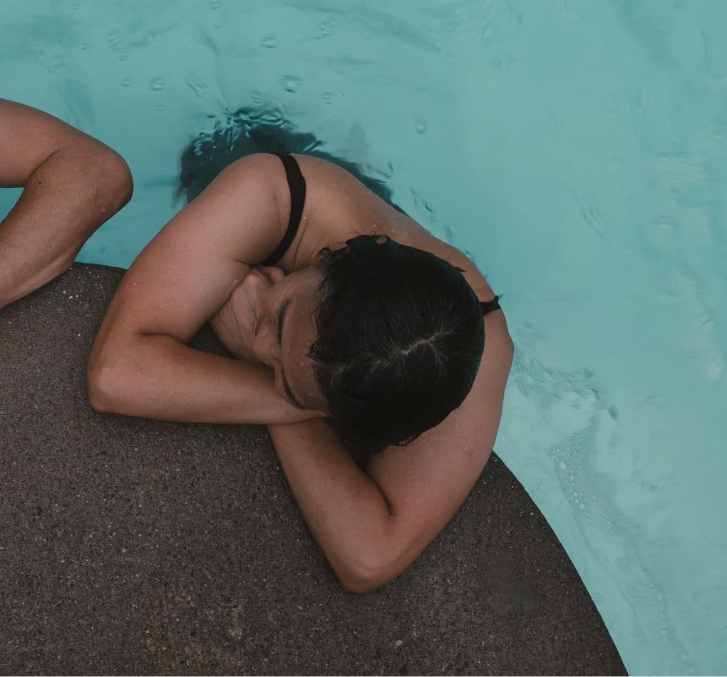 Woman relaxing beside her partner in a hot pool with clear blue water, while she leans forward on a smooth rock surface.